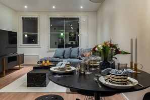 Un salon moderne avec une table à manger noire préparée pour quatre personnes, un canapé gris agrémenté de coussins à motifs et un téléviseur placé à gauche.