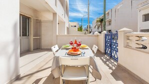 Un espace extérieur aménagé pour les repas, avec une table dressée pour deux personnes, des chaises blanches et une porte bleue en arrière-plan.