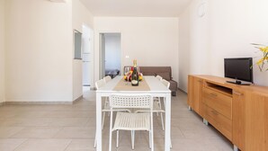 Une pièce moderne et bien éclairée, avec une table à manger dressée pour quatre personnes, un meuble en bois sur lequel est posée une télévision, et un canapé à l’arrière-plan.