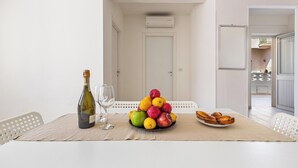 Un coin repas avec une table dressée pour deux personnes, agrémenté d’un panier de fruits, d’une bouteille de vin et d’une assiette de pâtisseries.