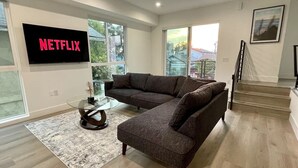 Una sala de estar moderna con un sofá modular, una mesa de centro de vidrio y un televisor de pantalla plana mostrando 'NETFLIX'.