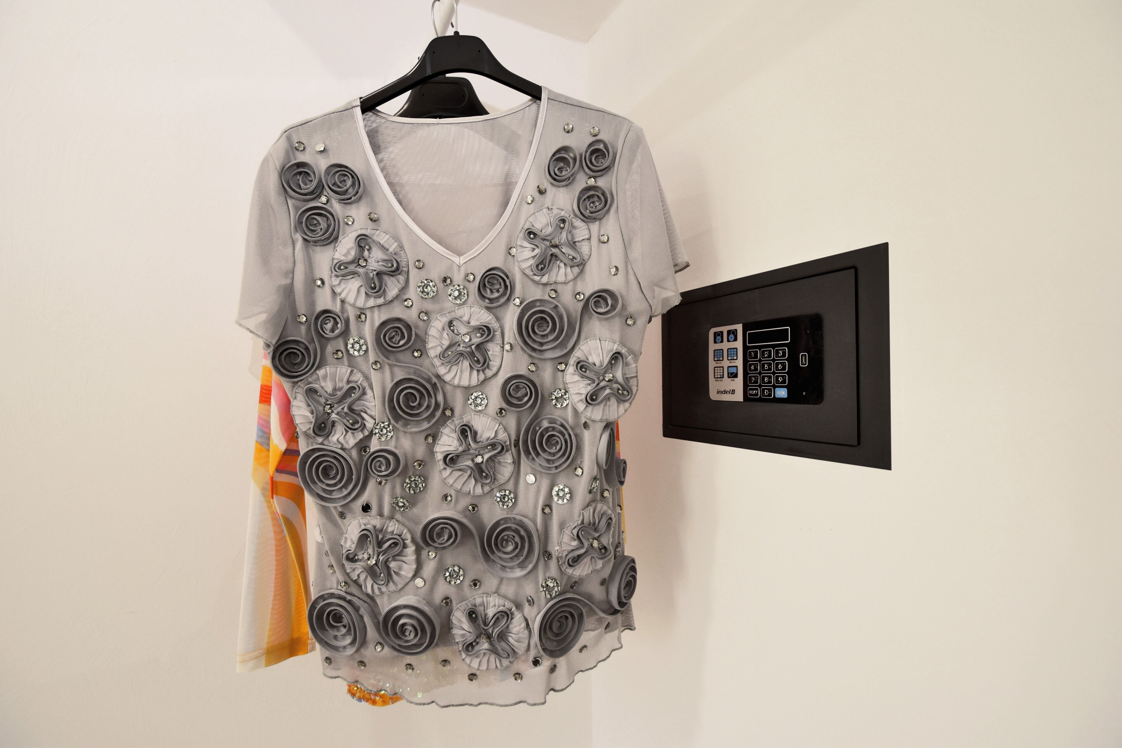 A garment with a V-neck and short sleeves, adorned with silver embellishments and buttons, hanging on a black hanger against a white wall.