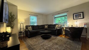 A modern living room with a black leather sofa, a round coffee table, and a patterned rug. There is a TV mounted on the wall, a floor lamp, and a desk with a chair.
