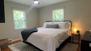 A bedroom with a bed, a ceiling fan, two windows with blinds, a bedside table with a lamp, and a small plant.