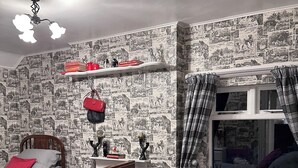 Une chambre avec deux lits, une tête de lit en bois, une table de chevet, une fenêtre avec des rideaux et un mur recouvert d’un papier peint à motifs.