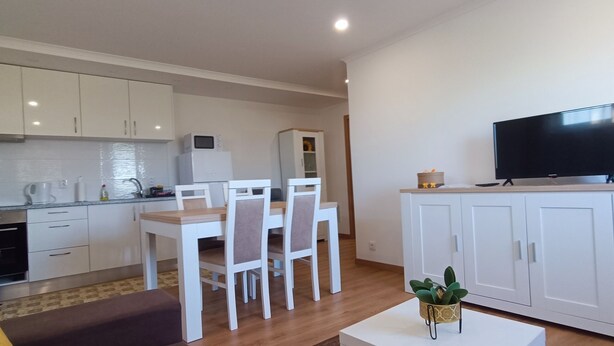 A modern kitchen with white cabinets, a dining area with a white table and chairs, a TV on a cabinet, and a small plant on a table. - Mondalva Apartments located in village of Quintela, near to São Pedro de Alva (São Pedro de Alva)