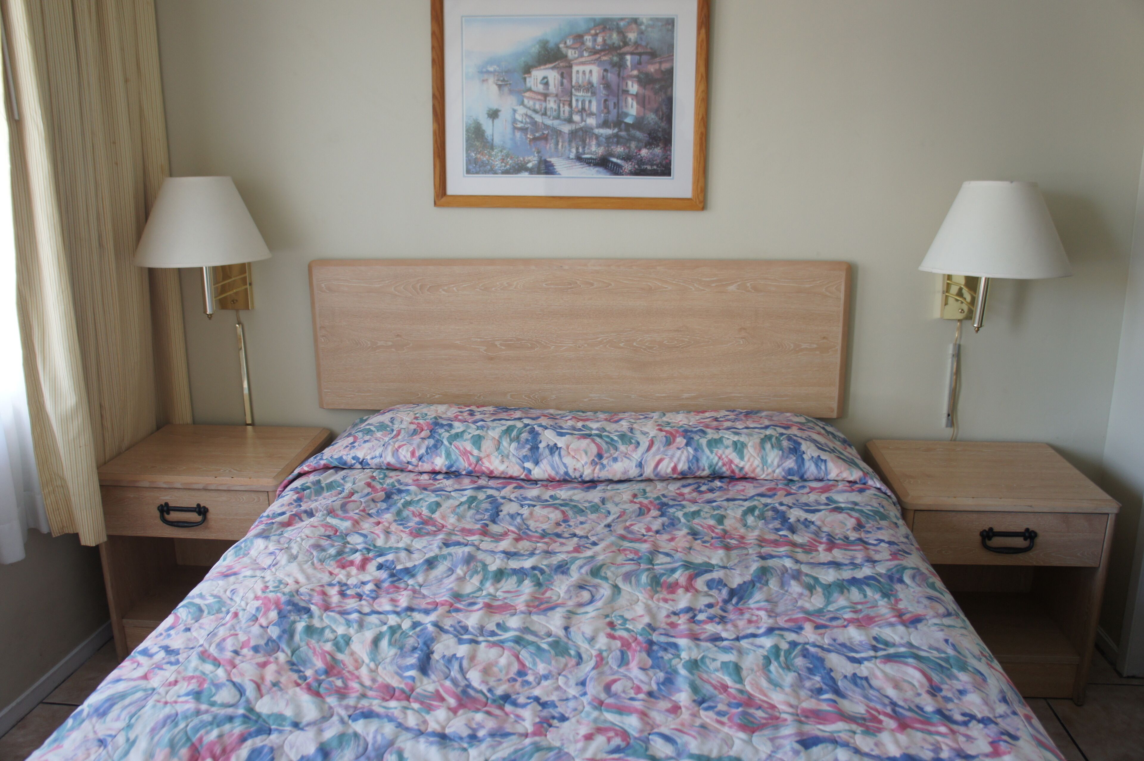 Un dormitorio con cabecera de madera, colcha con estampado floral, dos mesitas de noche con lámparas y un cuadro enmarcado en la pared.