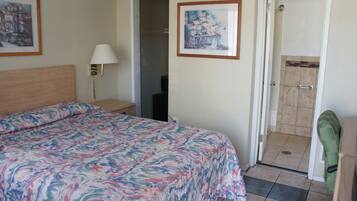 A hotel room with a bed, a desk, a chair, a lamp, and a shower area.