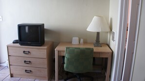 Um quarto pequeno com uma escrivaninha de madeira, uma cadeira verde, uma televisão em uma cômoda e um abajur.