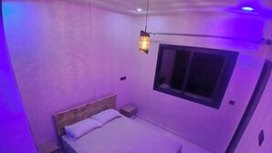 Une chambre avec un lit simple, une table de chevet, une commode, une fenêtre et un luminaire suspendu.