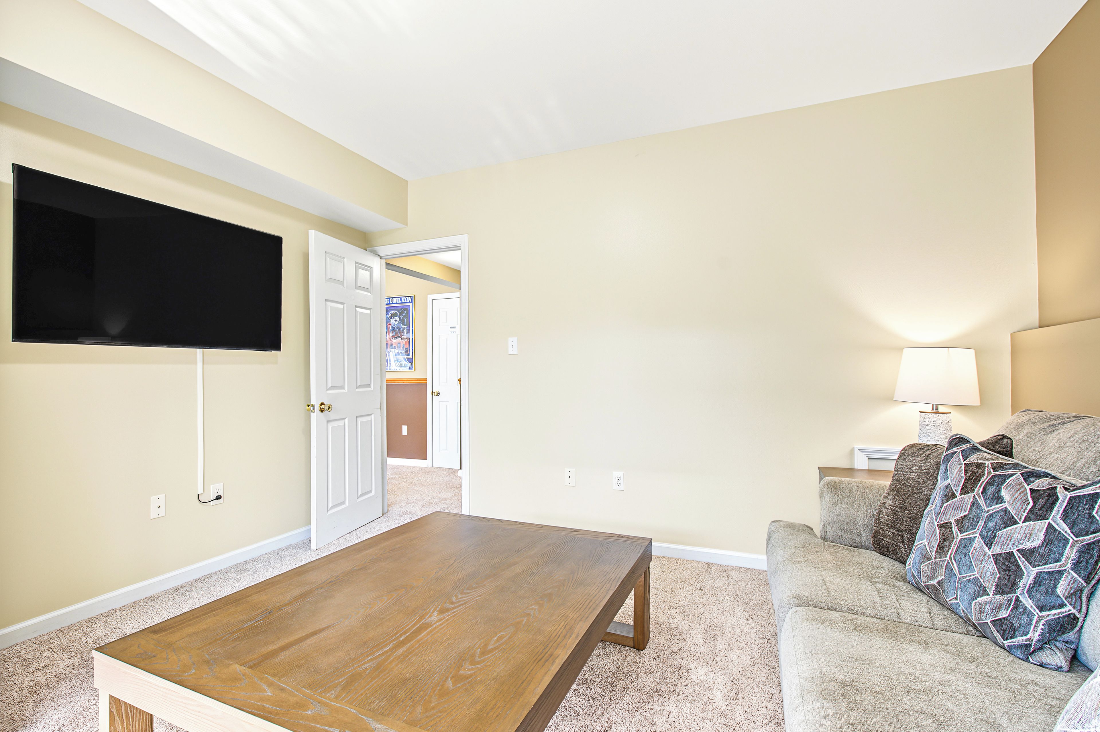A living room with a flat-screen TV mounted on the wall, a wooden coffee table, a patterned sofa, and a door leading to another room.