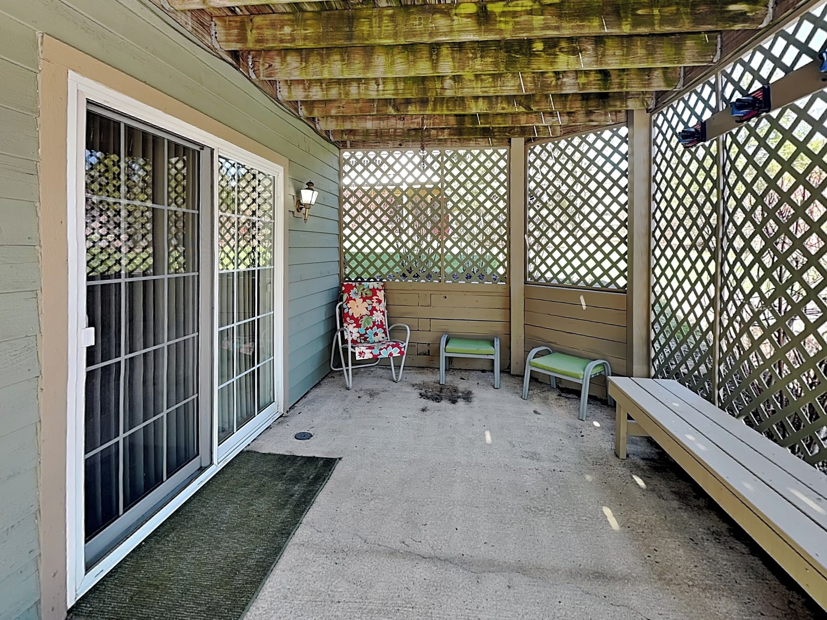 Un patio couvert avec une porte-fenêtre coulissante, un banc, une chaise et une clôture à treillis.