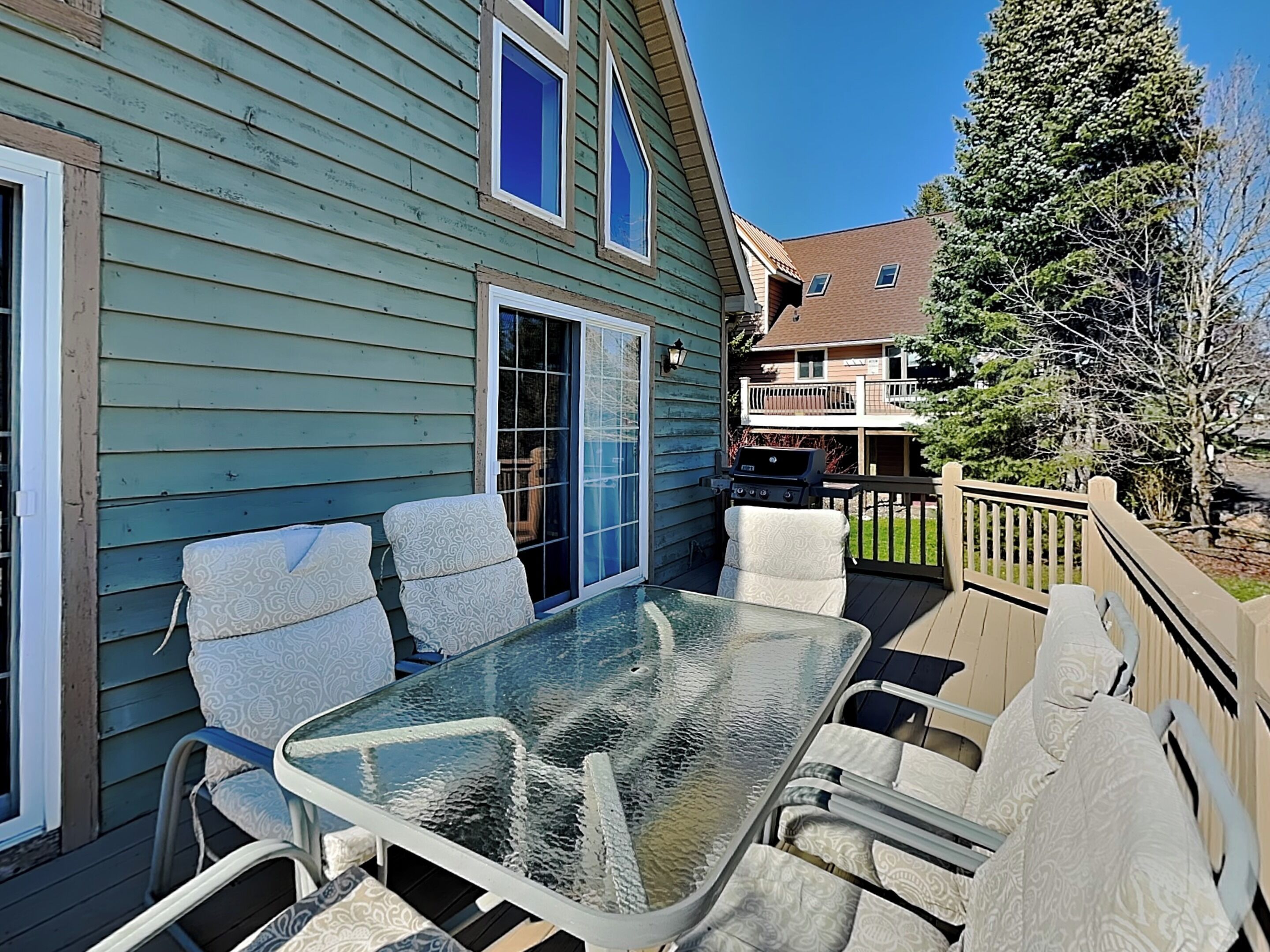 A glass-top table with chairs on a deck, a barbecue grill, and a house with a blue exterior.