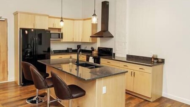 Una cocina moderna con armarios de madera, un refrigerador negro, un lavamanos de acero inoxidable y una barra con dos taburetes.