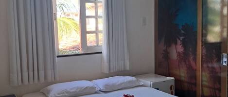 Habitación de hotel con cama, ventana con cortinas, aire acondicionado, mesita de noche y armario con un mural tropical.