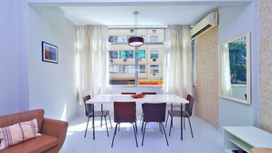 Een moderne woonkamer met een eethoek, met een bank, een eettafel, stoelen en een groot raam met gordijnen.