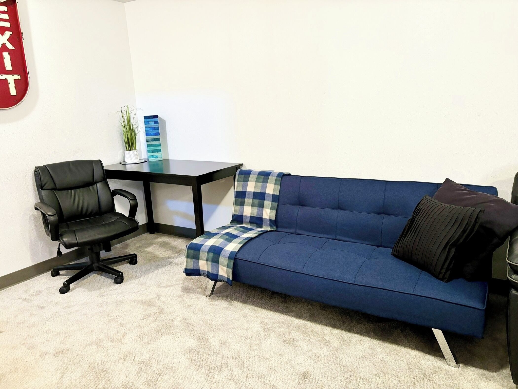 Um quarto com um sofá azul, uma cadeira de escritório preta, uma pequena mesa com uma planta e um sinal de saída vermelho na parede.