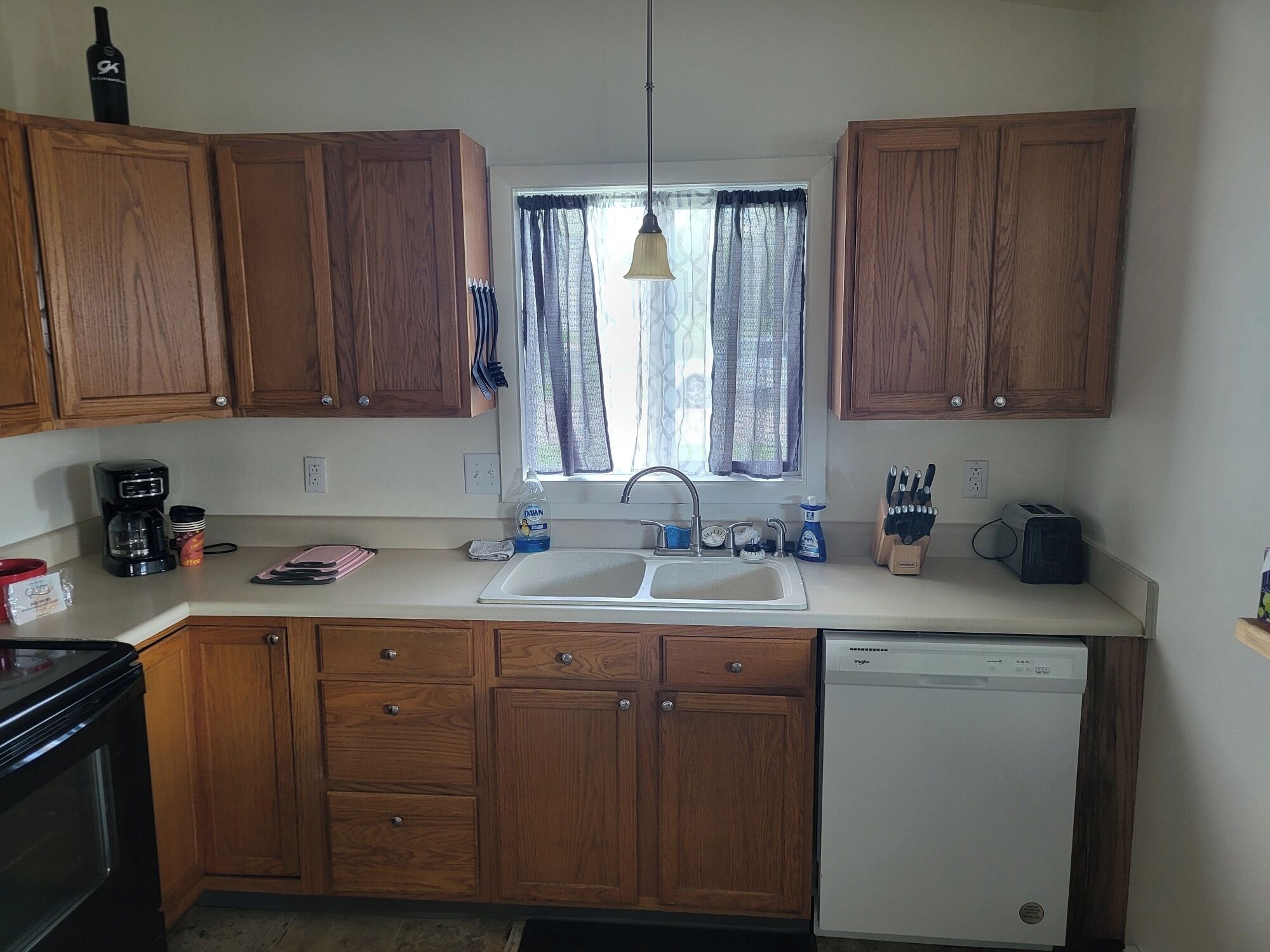 A kitchen with wooden cabinets, a dishwasher, a coffee maker, and a window with curtains.