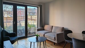 Una sala de estar moderna con un sofá gris, una mesita redonda y una silla negra. Unos ventanales correderos dan a un balcón con vistas a una valla de madera y edificios.