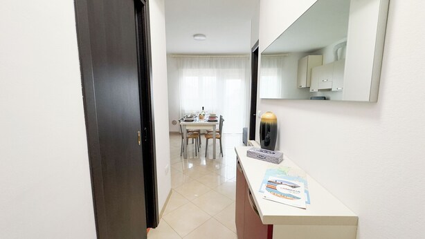 A hotel hallway with a dining area in the background, a white reception desk, and a large mirror on the wall. - Esperia Apartment by Ca-Sa Check (Chioggia)