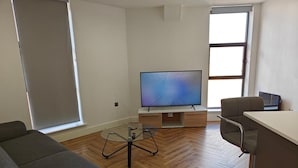 A modern living room with a flat-screen TV, a glass coffee table, a sofa, and a desk with a chair. - 2-bed Apartment Birmingham Snow Hill (Birmingham)