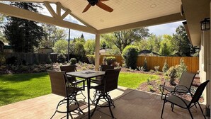 Un patio techado con una mesa y sillas, un ventilador de techo y vistas a un jardín trasero con cerca de madera y vegetación.