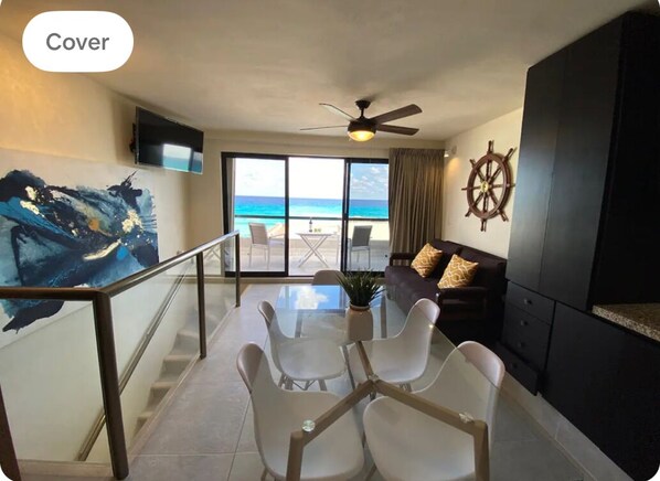 A modern living room with a glass table, white chairs, a ceiling fan, a TV, and a view of the ocean. - Luxury 2 beds apartment in front of the beach (Cancún)