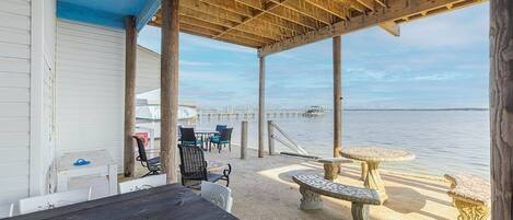 A covered outdoor seating area with tables and chairs, a stone bench, and a view of the water.