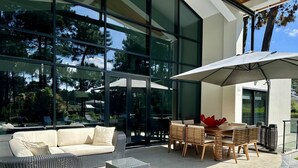 A modern outdoor seating area with a glass wall, a white sofa, wicker furniture, and a wooden table. - Quiet Villa with wonderful views of the golf courses! (Almada)