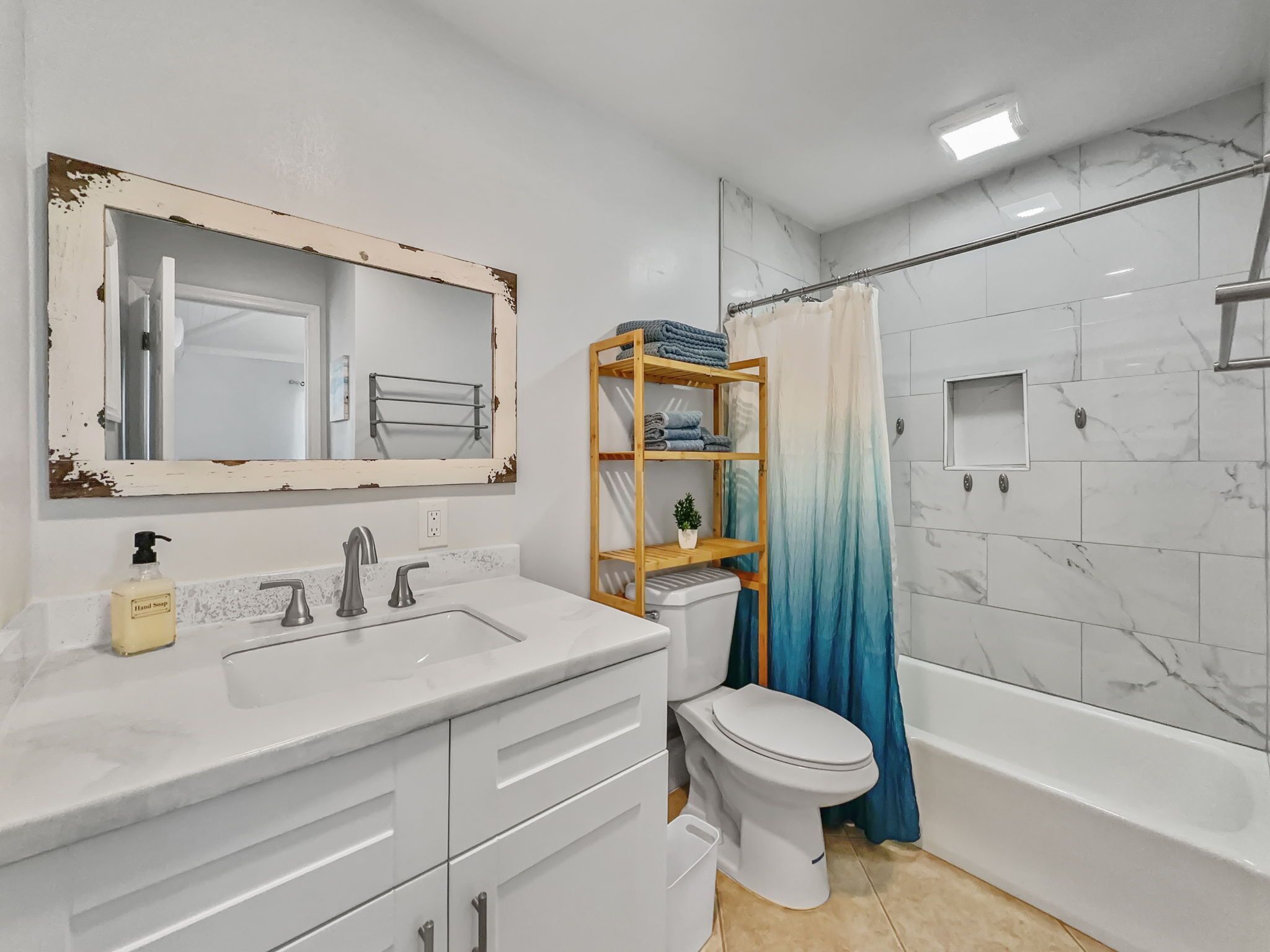 Une salle de bain avec un lavabo blanc, un miroir dont le cadre est vieilli, des toilettes, une étagère en bois avec des serviettes et un rideau de douche.
