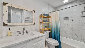 Une salle de bain avec un lavabo blanc, un miroir dont le cadre est vieilli, des toilettes, une étagère en bois avec des serviettes et un rideau de douche.