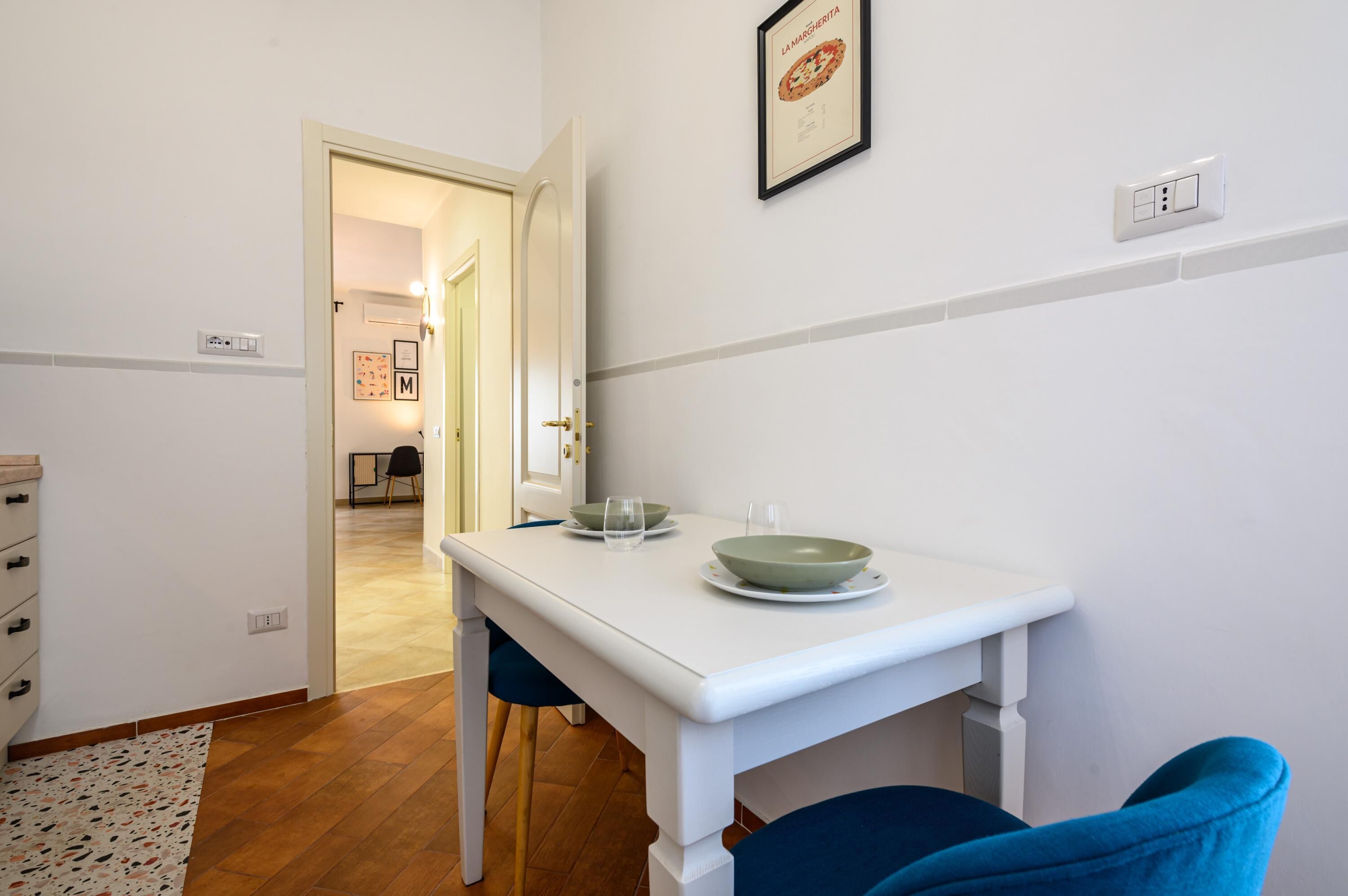 A clean, white room with a dining table set for two, a blue chair, and a door leading to another room.