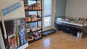 A kitchen with a refrigerator, shelves stocked with food items, a window with blinds, and a counter with various bottles. - Motel Claude (Degelis)
