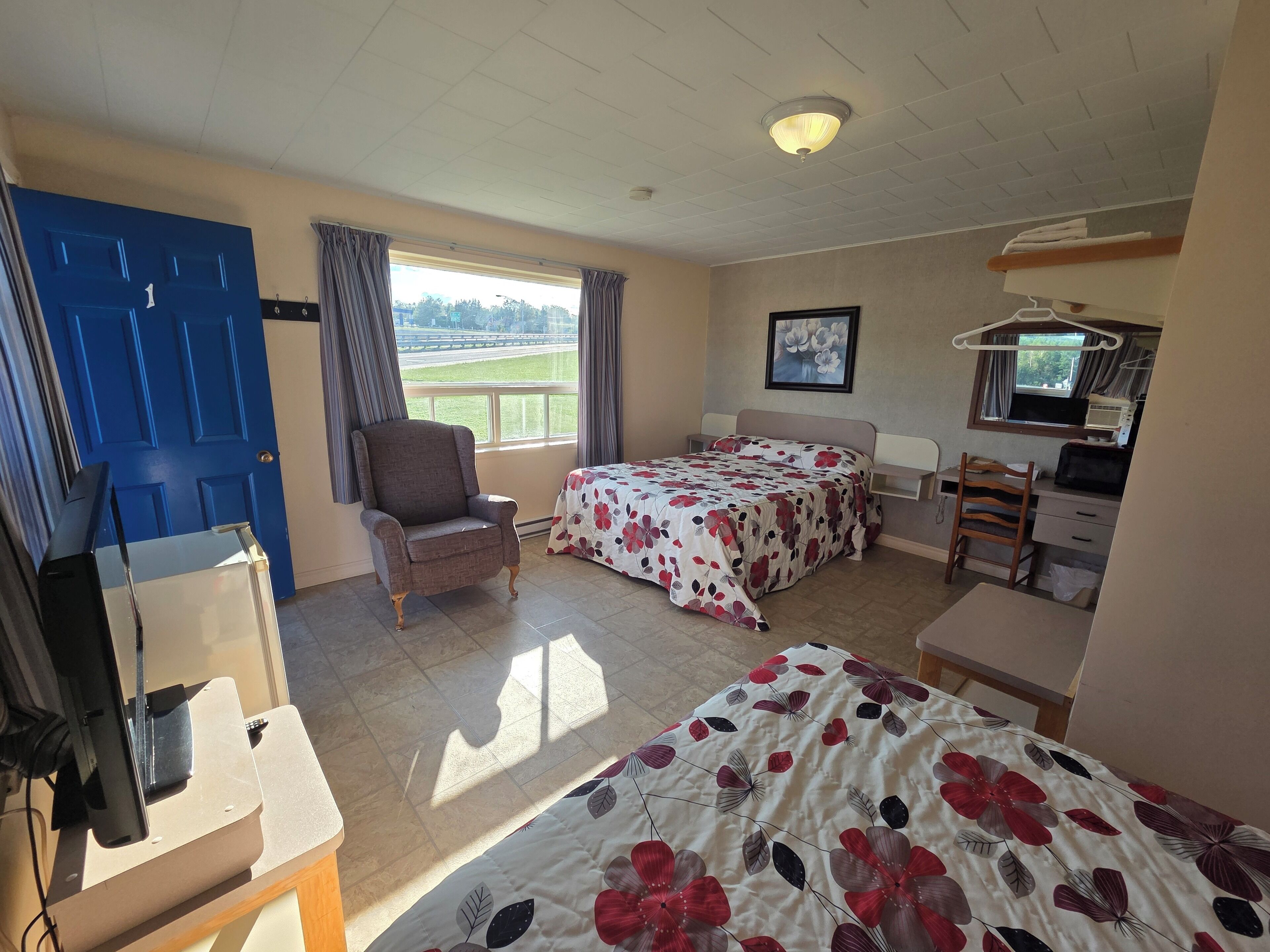 Una habitación con cama, una silla, un escritorio, televisor y ventana con cortinas.