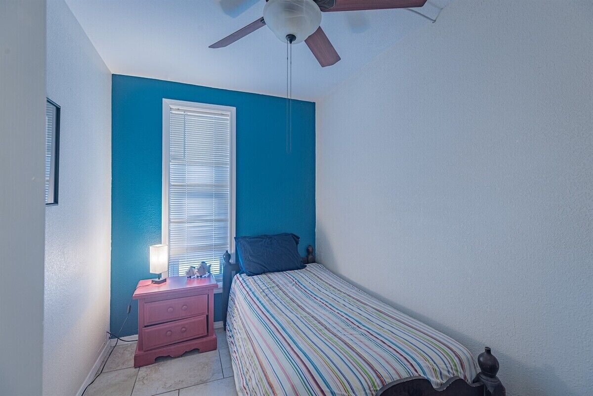Une chambre à coucher comprenant un lit, une table de chevet, une lampe, un ventilateur de plafond et une fenêtre avec des stores.