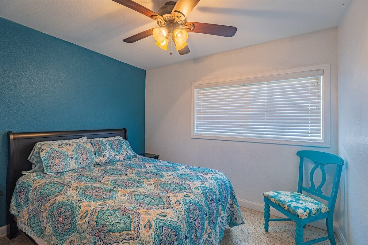 Une chambre à coucher avec un lit, un ventilateur de plafond, une fenêtre avec des stores et une chaise.