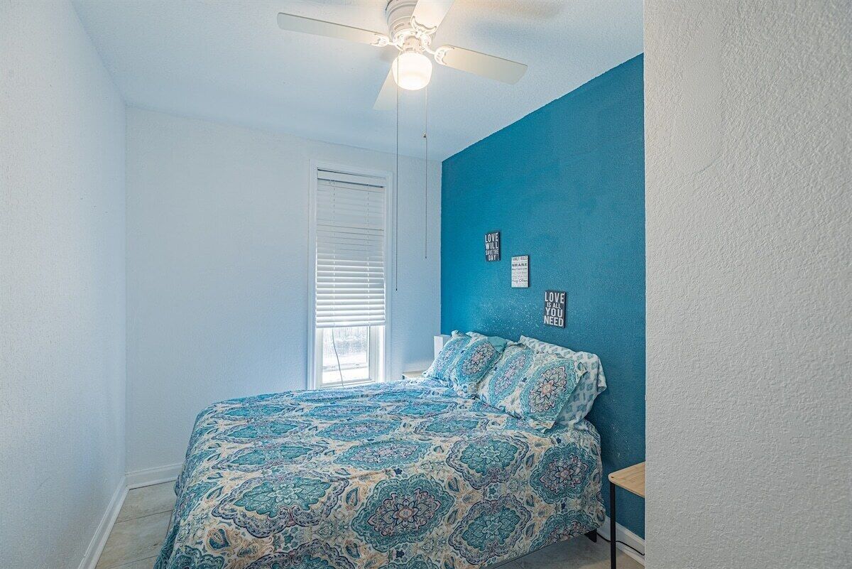 A bedroom with a bed, a ceiling fan, a window with blinds, and a blue wall with two framed motivational quotes.