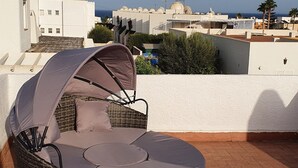 Un área de estar al aire libre con asientos de mimbre y un toldo, con vista a un pueblo costero con edificios y cielo despejado.