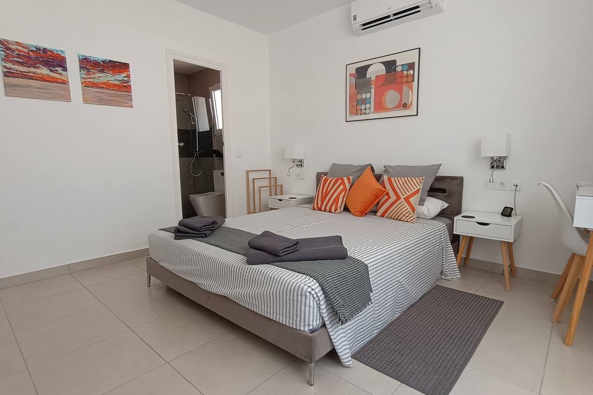 A modern bedroom with a bed, bedside tables, a desk, and a chair. There are two paintings on the wall, a shower area, and an air conditioning unit.