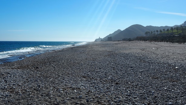 Una playa de arena con piedrecitas, cielo despejado y montañas a lo lejos.