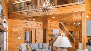 A spacious wooden cabin interior with a high ceiling, a central seating area with a sofa and armchairs, a dining table, and a staircase leading to an upper level. - Luxury Cabin centrally located to all the things to see and do in High Country! (Banner Elk)