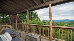 A wooden deck with outdoor furniture, including a sofa and a table, overlooking a forested landscape. - Luxury Cabin centrally located to all the things to see and do in High Country! (Banner Elk)
