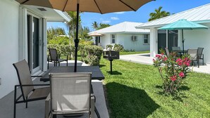A patio with a table and chairs, a large umbrella, and a small garden with pink flowers. - Golf Cart Included, Cat Island Villa, Treasure Cay, Bahamas (Treasure Cay)