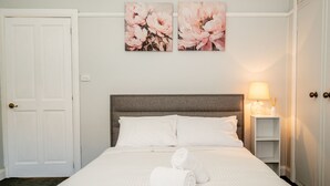 Habitación de hotel con cama, ropa de cama blanca, cabecero gris, mesita de noche con lámpara y dos cuadros florales en la pared.