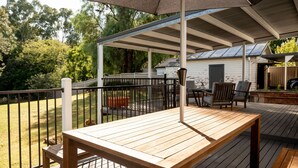 Una terraza de madera con mesa y sillas, un espacio cubierto con techo, y un edificio blanco al fondo.