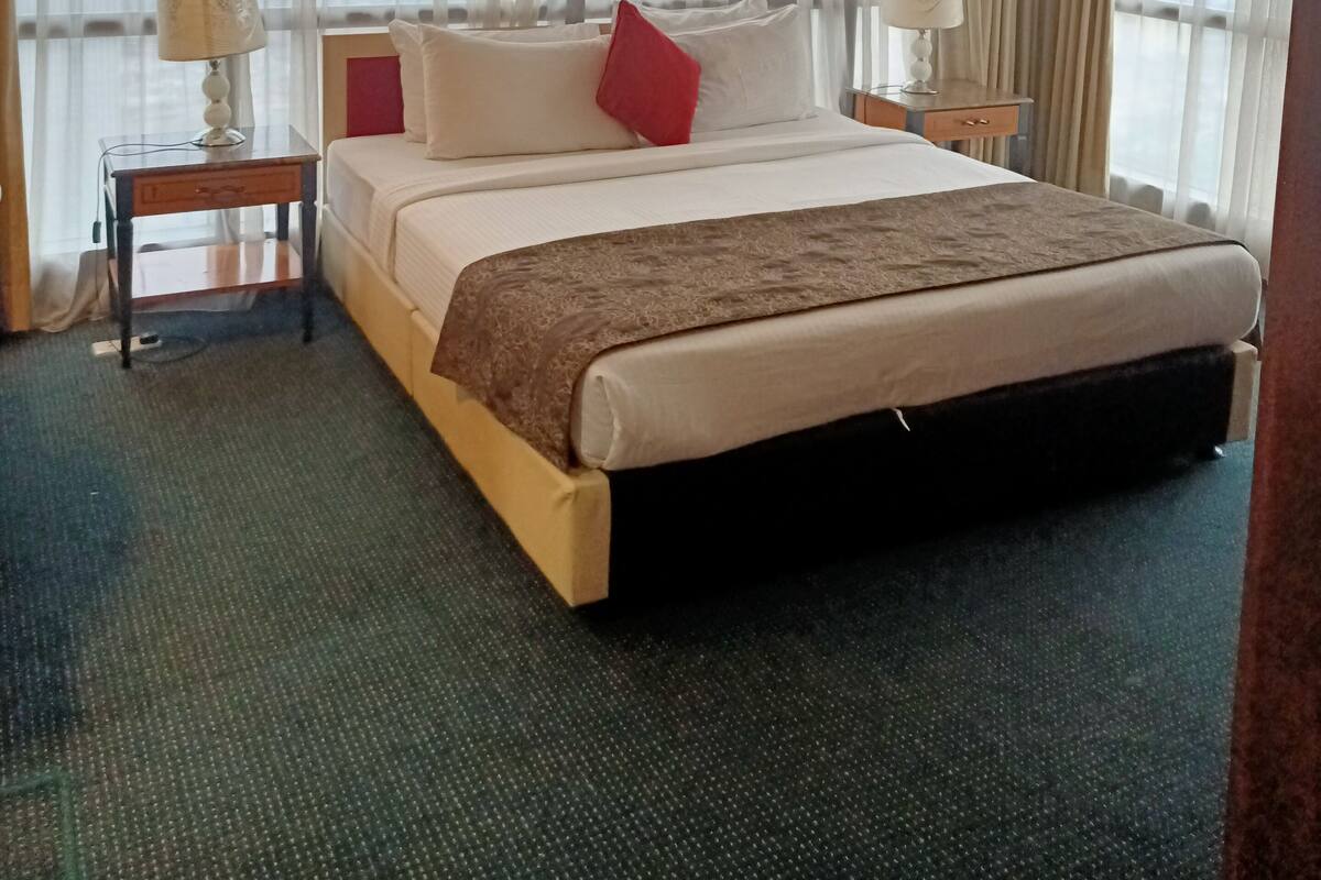 Ein Hotelzimmer mit einem großen Bett, zwei Nachttischen mit Lampen, durchsichtigen Vorhängen und Blick auf die Stadtlandschaft.