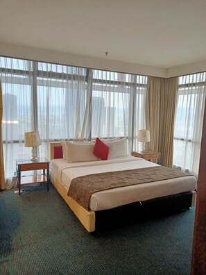 A hotel room with a large bed, two bedside tables with lamps, sheer curtains, and a view of the cityscape.