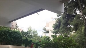An outdoor patio with a glass table and black metal chairs, surrounded by greenery and a view of buildings.