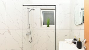 Un bagno moderno con doccia vetrata, un piccolo oblò con una pianta e un lavandino bianco con dispenser di sapone neri.
