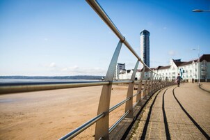 A coastal promenade with a metal railing, a sandy beach, and buildings in the background.